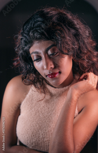 A woman with dark hair and a tan shirt is sitting with her arm around her neck. She has red lipstick on and is looking at the camera