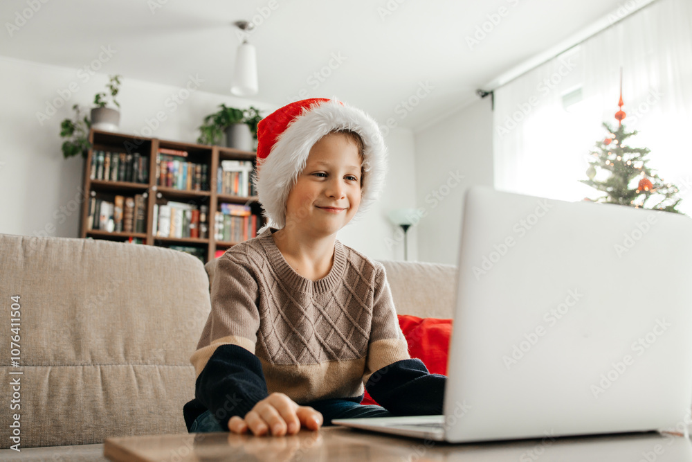 Happy boy in santa hat sitting on sofa in front of laptop. A child congratulates relatives online on Christmas Eve Happy boy in santa hat sitting on sofa in front of laptop. A child congratulates relatives online on Christmas Eve