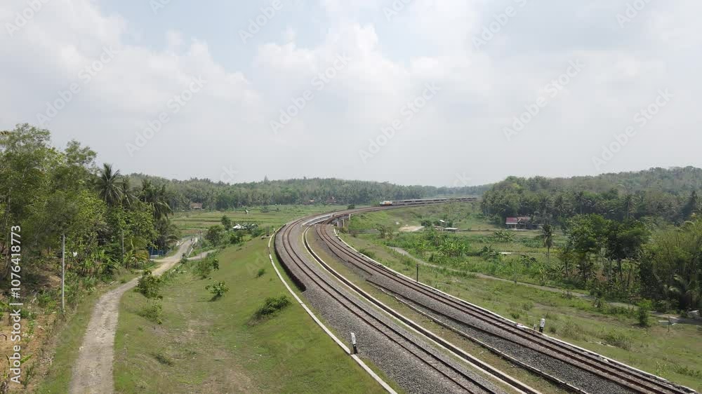 Drone shot, Indonesian train passing through a double track railway bridge winding on the left and right with rice fields, forests and villages.