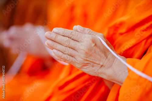 Close-up of a Buddhist monk's hands in a prayer gesture, holding a white string during a traditional blessing ceremony. The orange robes symbolize peace and devotion in Buddhist culture