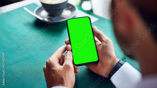 A mockup image of a business person holding a smart mobile phone with a blank green screen during a meeting or lunch in a modern cafe restaurant.