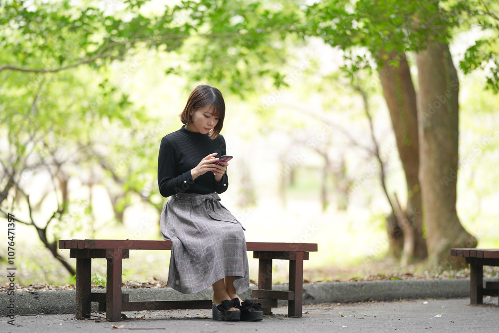 A young woman in her 20s with short hair wearing a black knitted long-sleeved shirt is sitting on a bench in the forest and operating a smartphone. A young woman in her 20s with short hair wearing a black knitted long-sleeved shirt is sitting on a bench in the forest and operating a smartphone.