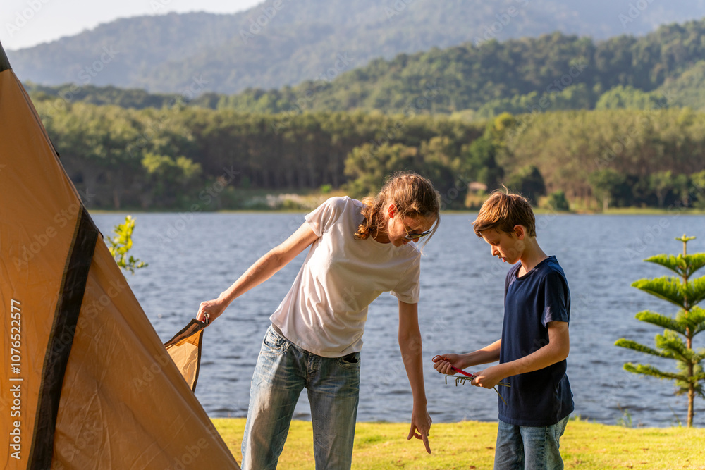 Fototapeta premium Mother and young son boy setting up a large, beige camping tent on a grassy area near a lake work together shared camping experience feelings of relaxation for a camping or outdoor adventure concept.