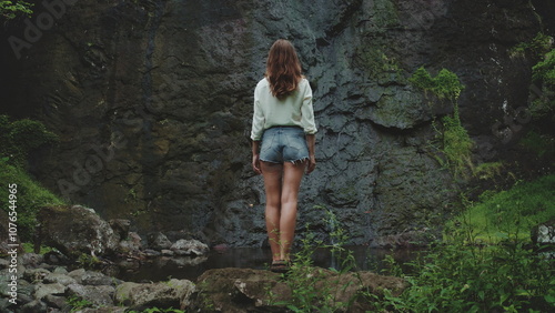 A young woman in a white shirt and denim shorts stands on a rocky outcrop near a waterfall in a lush, tropical forest in French Polynesia. The waterfall cascades down a dark, mossy rock face.