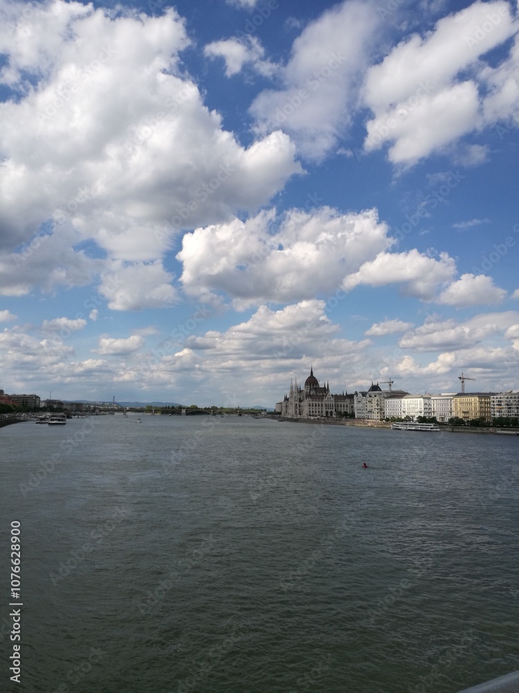 Naklejka premium A peaceful river view under a blue sky with scattered clouds and historic buildings along the bank in an urban setting
