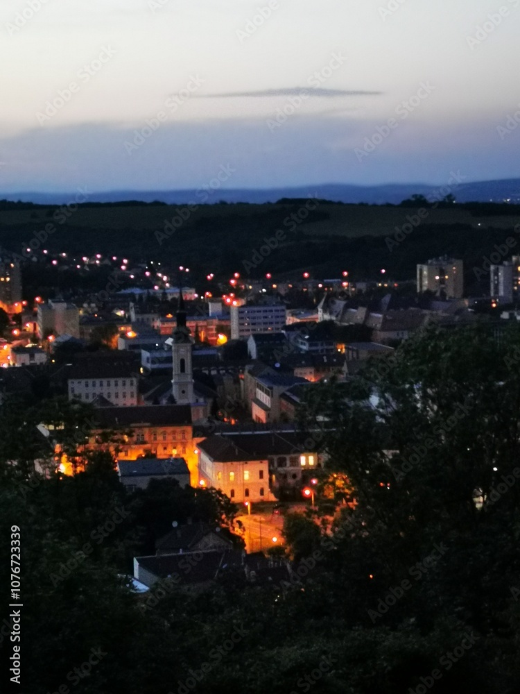 Fototapeta premium View of a warm-lit historic town under evening sky with distant hills and twinkling lights, captured during twilight hours