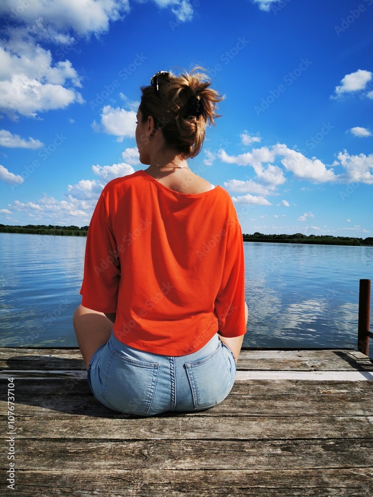 A woman enjoys a serene moment by the lake while sitting on a wooden dock under a bright blue sky with fluffy clouds A woman enjoys a serene moment by the lake while sitting on a wooden dock under a bright blue sky with fluffy clouds