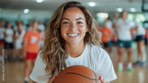 A cheerful young woman stands with a basketball in a busy indoor sporting environment, conveying excitement and camaraderie, attracting focus on teamwork and joy. photo