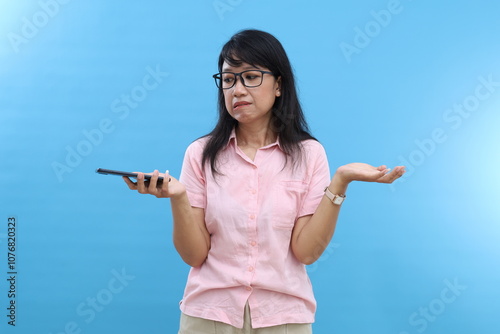 Photo of confused dark haired with asian appearance, holds mobile phone, spreads hands sideways, bites lower lip confusingly, cannot understand how download application, wears casual clothes photo