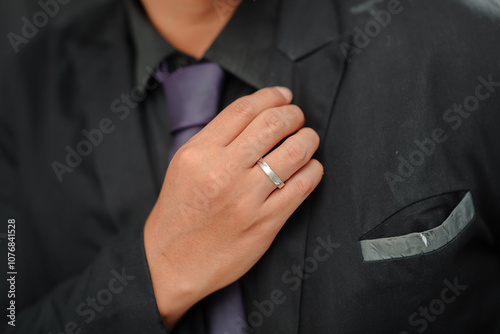 Close-up of a man in a black suit adjusting his collar, wearing a silver wedding band on his ring finger. The suit is paired with a dark tie, conveying a formal and elegant look.