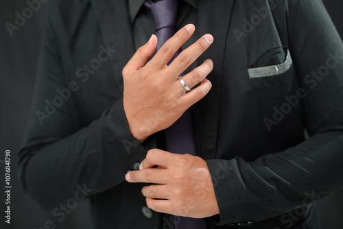 Close-up of a man in a black suit adjusting his collar, wearing a silver wedding band on his ring finger. The suit is paired with a dark tie, conveying a formal and elegant look.