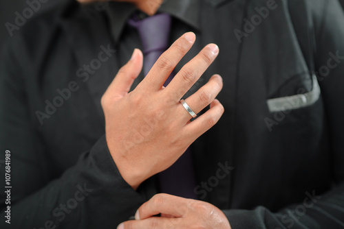 Close-up of a man in a black suit adjusting his collar, wearing a silver wedding band on his ring finger. The suit is paired with a dark tie, conveying a formal and elegant look.