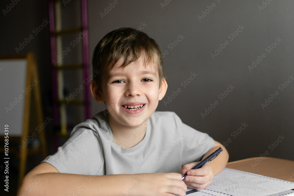 A boy with blond hair in a white preschool T-shirt sits at a table in his room and writes numbers in a notebook with his left hand A boy with blond hair in a white preschool T-shirt sits at a table in his room and writes numbers in a notebook with his left hand