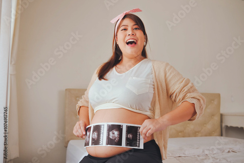 Joyful pregnant woman holding ultrasound photos over her belly, smiling widely, and expressing excitement about her pregnancy, wearing a pink headband and casual clothes. photo