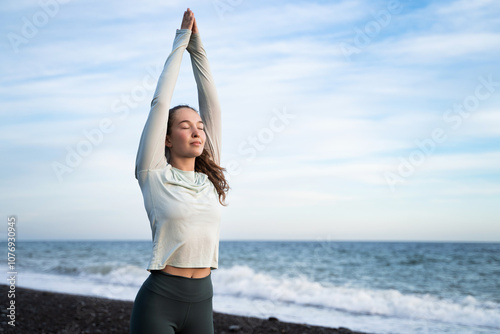 Fototapeta Naklejka Na Ścianę i Meble -  Peaceful young woman meditating on seashore at sunrise, sporty girl stretching arms up by the sea and sky, breathing fresh air and doing yoga on beach, morning run, health wellbeing sport activities