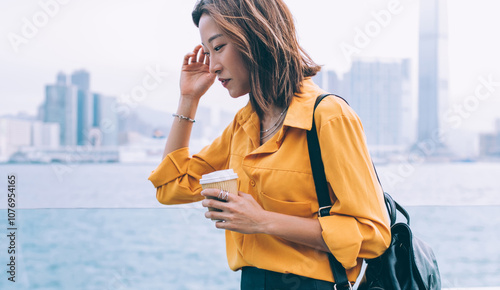 Thoughtful female tourist with coffee to go in hand pondering on vacations near river in Hong Kong, attractive hipster woman trendy dressed thinking on discover metropolis during Chinese holidays #1076954165