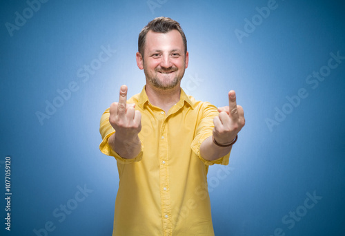 Young man showing fuck off gesture over blue background, dresses in yellow shirt. Male person with obscene gesture giving the middle finger and looking to camera photo