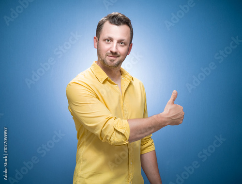 Man with thumbs up, like concept over blue background, dresses in yellow shirt. Yes gesturing, sings of good work. Half length of pleased guy photo