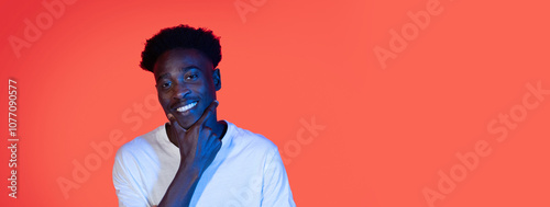 African American young man poses confidently with a slight smile, hand thoughtfully resting on his chin. The bright orange backdrop adds a lively touch, enhancing the overall cheerful vibe. photo