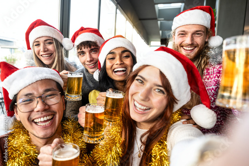 Cheerful young people sitting celebrating x-mas eve drinking beer at brewery pub restaurant - Happy friends wearing santa claus hat having Christmas dinner party - Winter holidays portrait concept