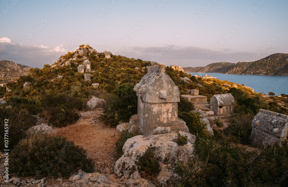 Fototapeta premium Stone sarcophagus from ancient Simena Kaleköy city in Turkey. Mediterranean sea surrounded by rugged hills listed morning light. Archaeological treasure of Lycia.