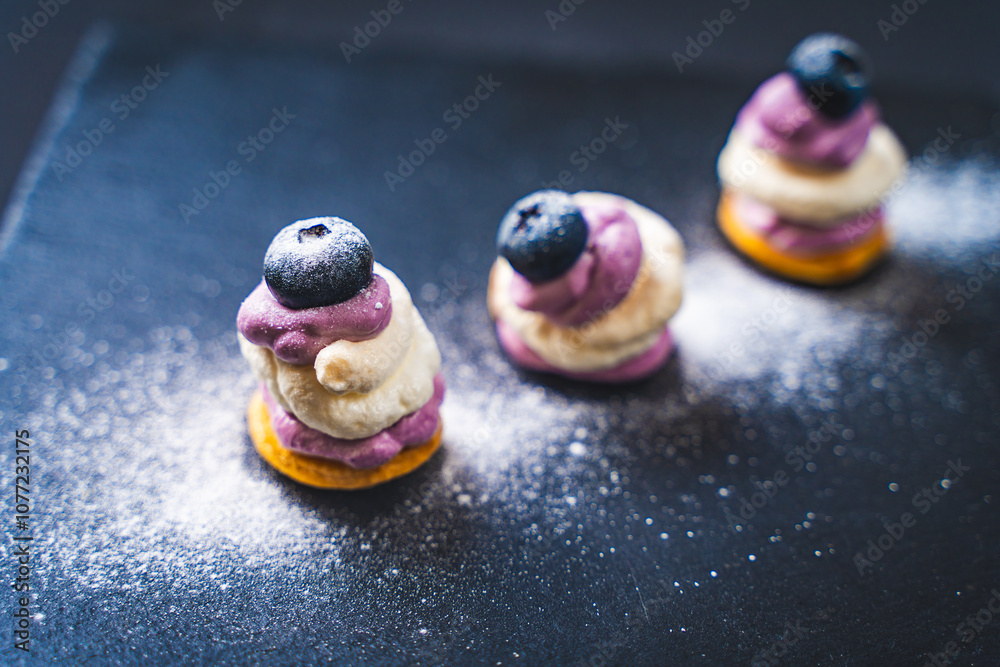Fototapeta premium The cake lost its shape next to the two well-standing. Mini desserts on a black mica board with blueberry cream and blueberries, sprinkled with powdered sugar