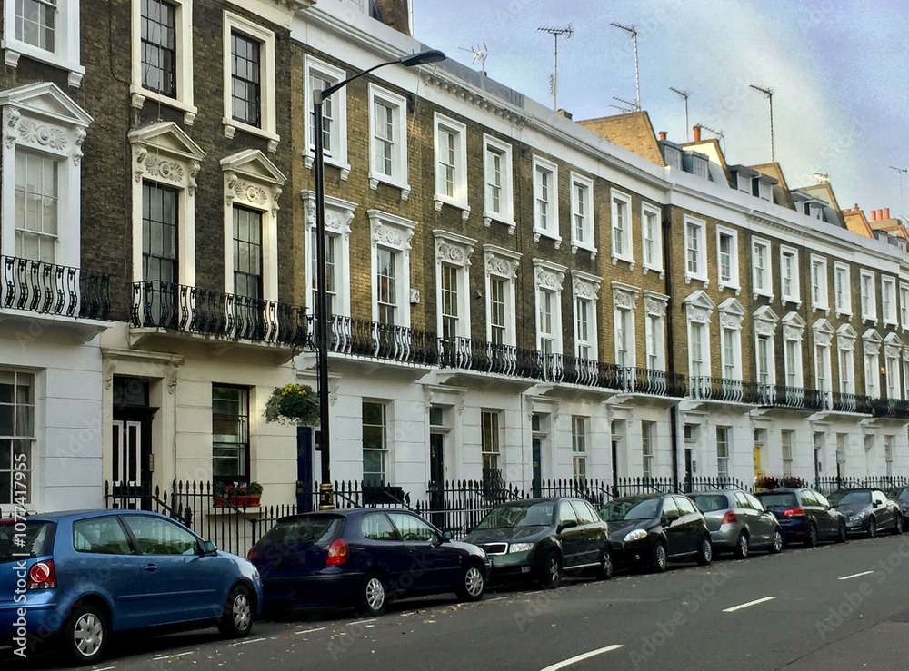 Fototapeta premium A row of traditional English townhouses with intricate architectural details, alongside parked cars on a quiet street. The image showcases a classic urban residential scene.
