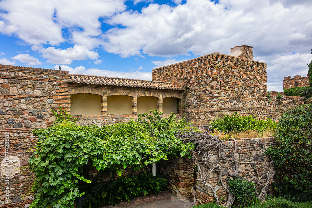 A view of Malaga's Alcazaba walls, featuring historic stone walls, defensive towers, lush greenery, and a blue sky. Impressive medieval Moorish architecture. Andalusia, Spain. A view of Malaga's Alcazaba walls, featuring historic stone walls, defensive towers, lush greenery, and a blue sky. Impressive medieval Moorish architecture. Andalusia, Spain.