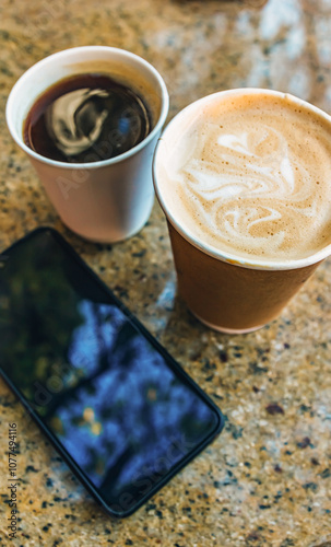 Two paper coffee cups with americano and cappuccino on a stone table at an outdoor cafe. A smartphone lies nearby, adding a modern touch to the cozy street setting