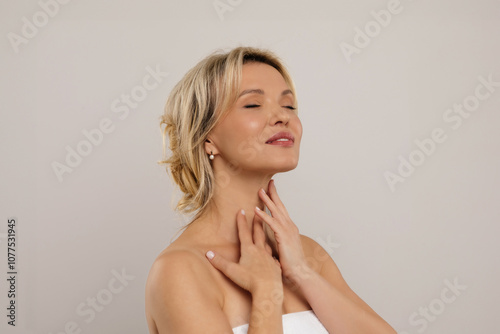 Happy smiling attractive half-naked middle aged blonde woman covered in towel touching her neck, enjoying anti-aging SPA salon treatment results, grey studio background, copy space. High quality photo