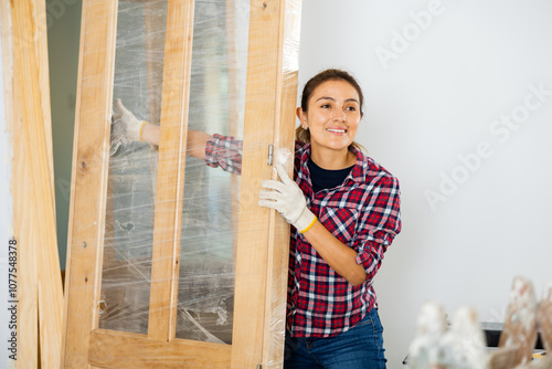 Smiling latin woman working in construction site in apartment, carrying new wooden door.