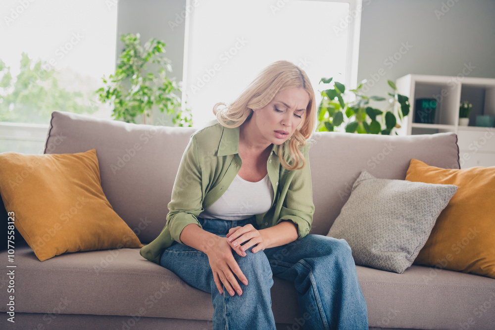 Middle-aged woman with blond hair sitting on sofa in bright living room, feeling discomfort and pain. Middle-aged woman with blond hair sitting on sofa in bright living room, feeling discomfort and pain.
