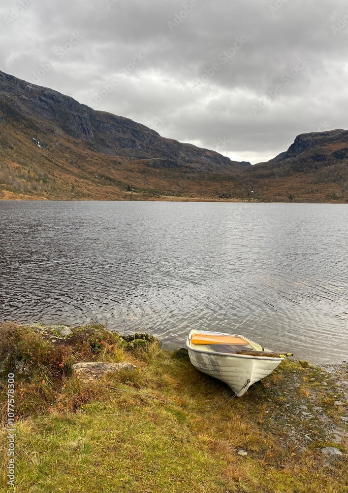 A peaceful image of a tranquil lake surrounded by rugged mountains, with an abandoned rowboat on the grassy shore, under a cloudy sky. Solitude and nature in the fall in the west of Norway. A peaceful image of a tranquil lake surrounded by rugged mountains, with an abandoned rowboat on the grassy shore, under a cloudy sky. Solitude and nature in the fall in the west of Norway.