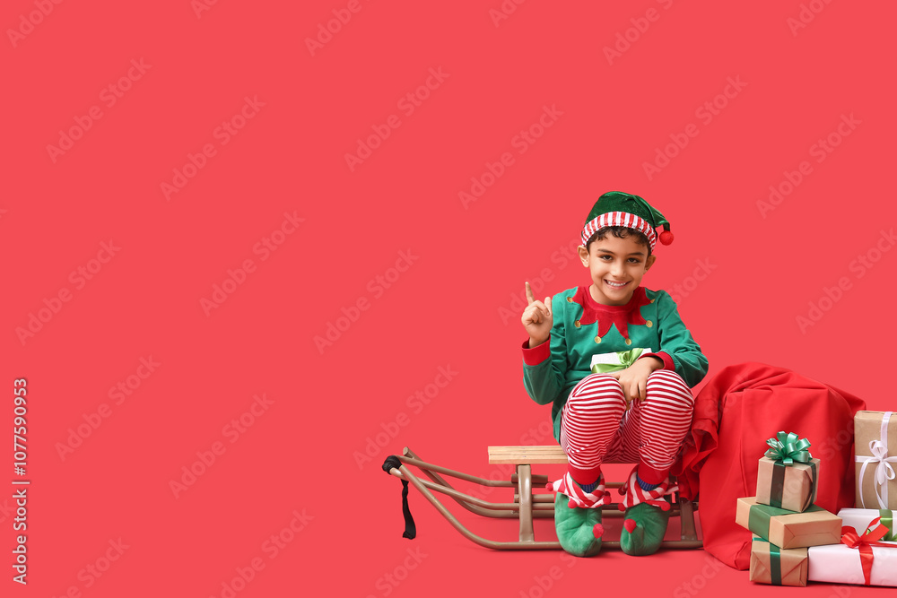 Naklejka premium Cute little elf with sled and Christmas gifts pointing at something on red background