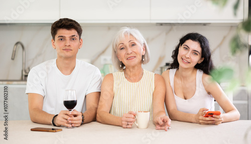 Elderly woman is sitting at table between young people, guy and girl, smiling sweetly and posing