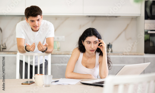 Young guy screams during family quarrel with young woman using laptop in kitchen