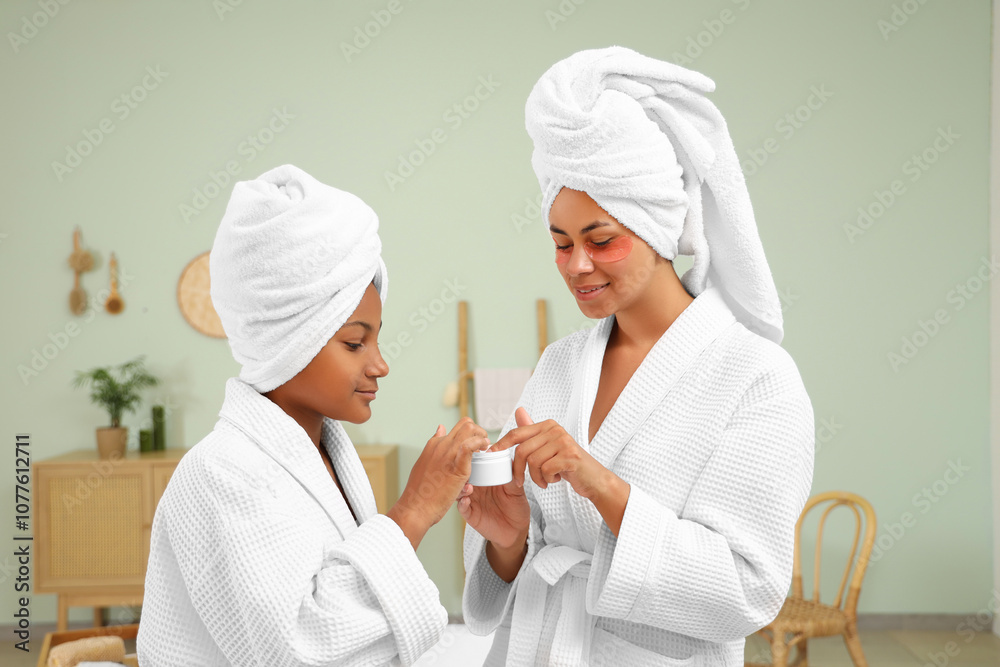 Happy young African-American mother and her daughter in bathrobes with under-eye patches applying facial cream at home Happy young African-American mother and her daughter in bathrobes with under-eye patches applying facial cream at home