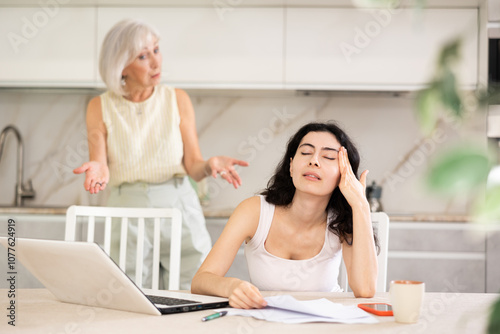 Elderly woman screaming during family quarrel with young woman using laptop in kitchen