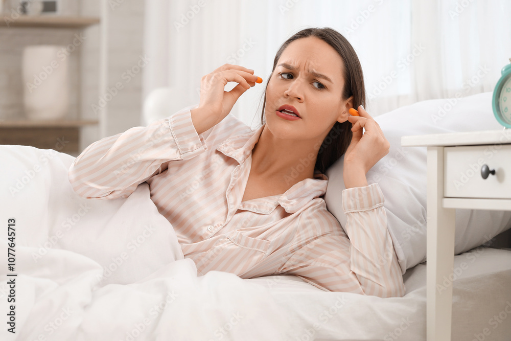 Displeased young woman with ear plugs lying in bedroom Displeased young woman with ear plugs lying in bedroom