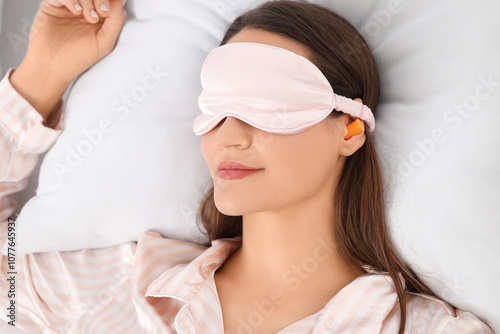 Young woman with sleeping mask and ear plugs lying in bedroom, closeup photo