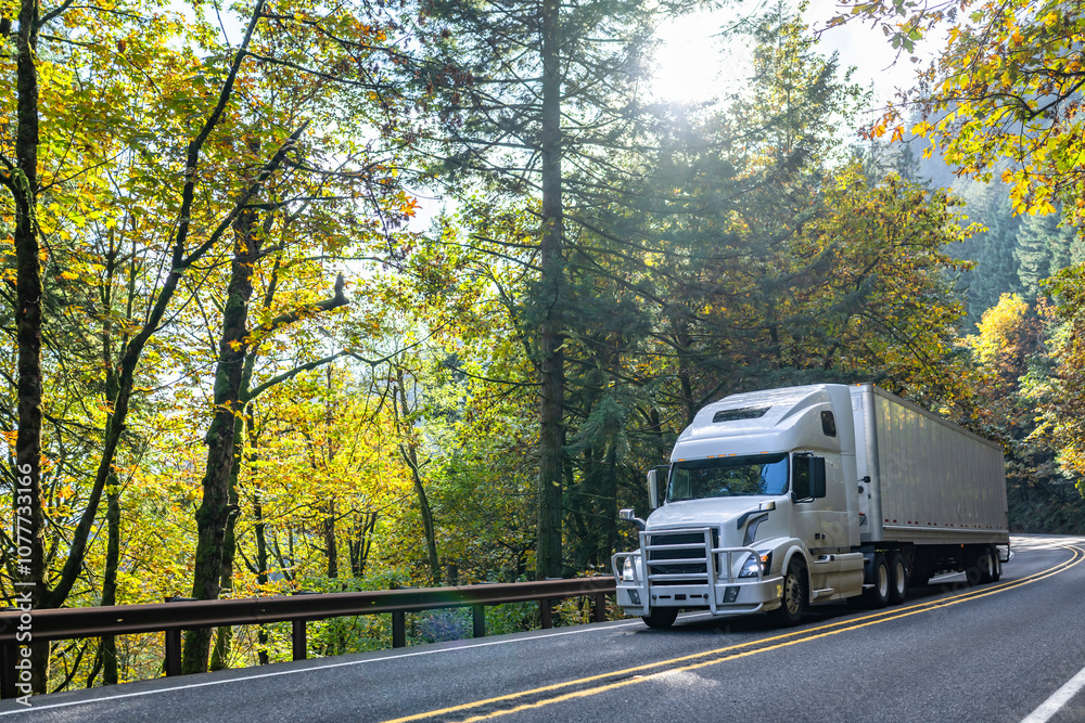 Popular white big rig semi truck with grille guard transporting cargo in dry van semi trailer running on the winding autumn mountain road with forest on the sides at sunlight Popular white big rig semi truck with grille guard transporting cargo in dry van semi trailer running on the winding autumn mountain road with forest on the sides at sunlight