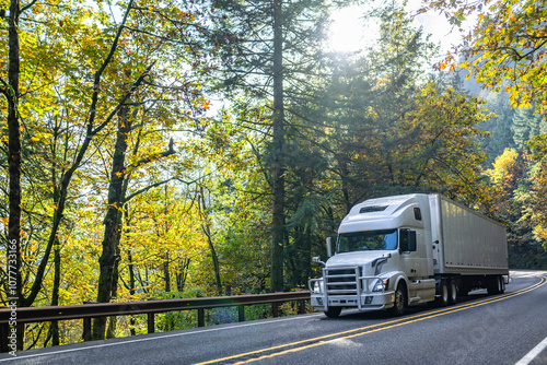 Popular white big rig semi truck with grille guard transporting cargo in dry van semi trailer running on the winding autumn mountain road with forest on the sides at sunlight