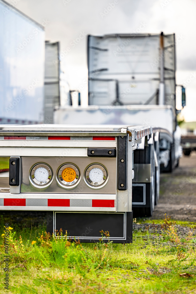 White big rig semi truck with empty step deck semi trailer with round rear lights standing on the parking lot with green grass White big rig semi truck with empty step deck semi trailer with round rear lights standing on the parking lot with green grass