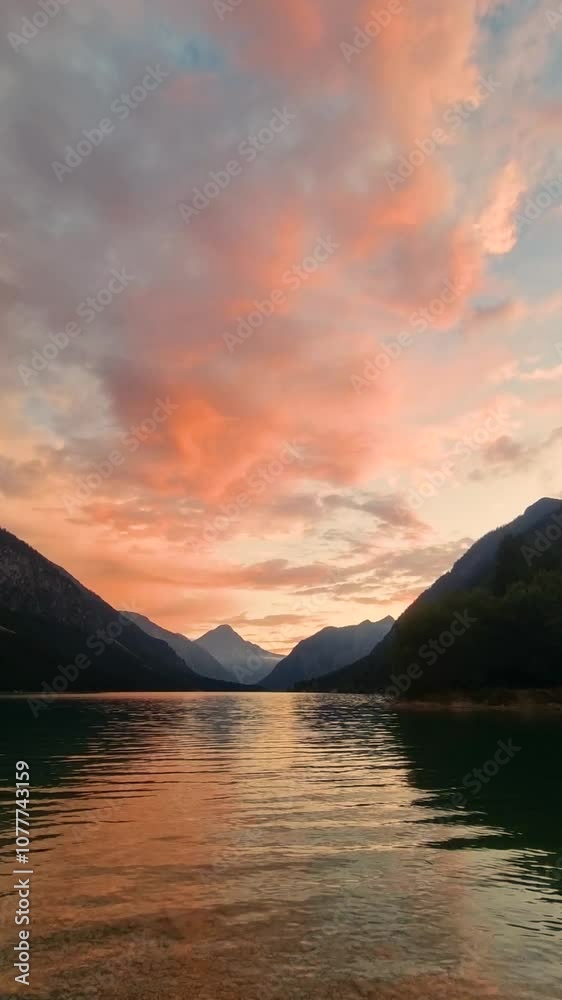 The sky transforms into a canvas of orange and pink hues as the sun sets calmly over a serene lake. Majestic mountains mirror the reflective waters, creating a picturesque landscape. Plansee Austria The sky transforms into a canvas of orange and pink hues as the sun sets calmly over a serene lake. Majestic mountains mirror the reflective waters, creating a picturesque landscape. Plansee Austria