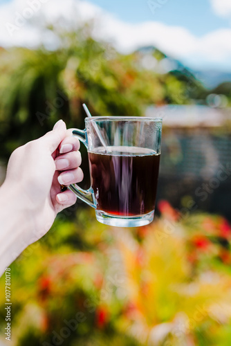 hand holding coffee in clear mug in front of blurred home interior background, habits and lifestyle concept photo