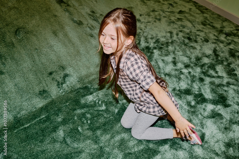 Cheerful little girl in cotton shirt and grey leggins sitting on the floor next to soft green slide after rolling down during play Cheerful little girl in cotton shirt and grey leggins sitting on the floor next to soft green slide after rolling down during play