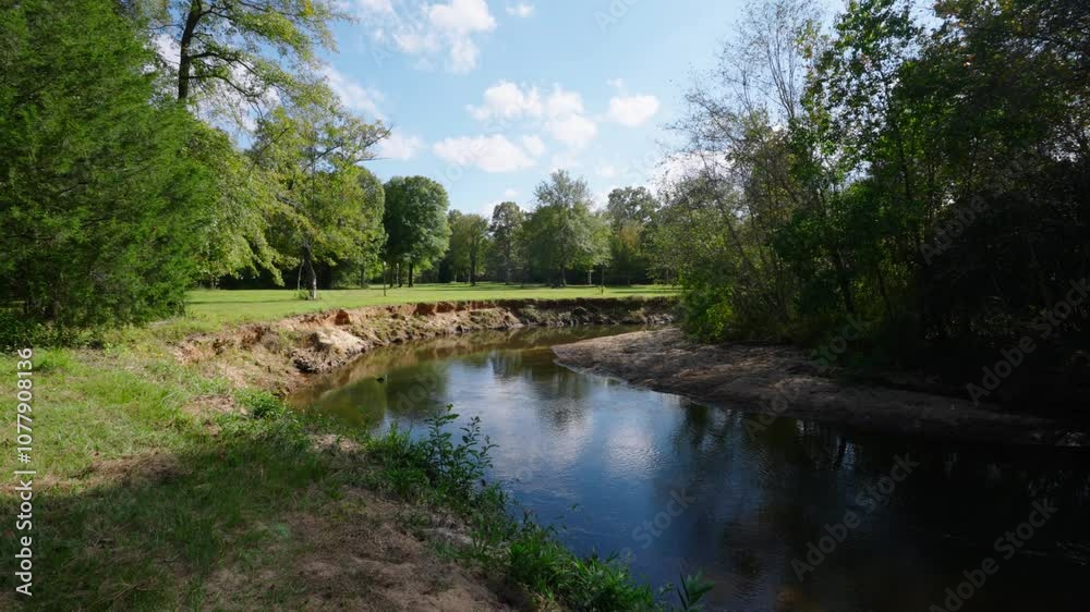 A scenic river flowing through a grassy area with tall, eroded banks and surrounded by lush greenery and trees. The sunny day and clear sky add a peaceful, rural charm to the natural landscape. A scenic river flowing through a grassy area with tall, eroded banks and surrounded by lush greenery and trees. The sunny day and clear sky add a peaceful, rural charm to the natural landscape.
