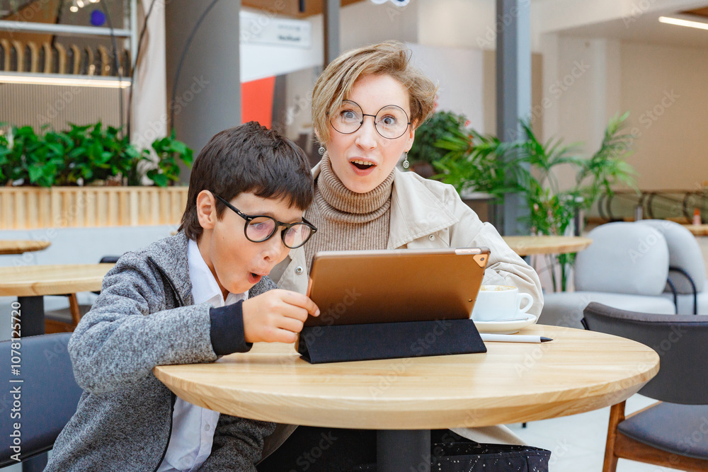 A young boy and a woman both are in glasses, looking at a tablet together, using wireless technology and communication A young boy and a woman both are in glasses, looking at a tablet together, using wireless technology and communication