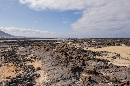 White sand beach with solidified volcanic lava flows, black rocks. Mountains in the background and sky with big white clouds. Caletón Blanco, Lanzarote, Canary Islands, Spain