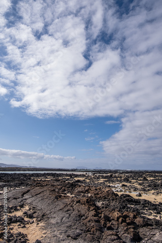 White sand beach with solidified volcanic lava flows, black rocks. Mountains in the background and sky with big white clouds. Caletón Blanco, Lanzarote, Canary Islands, Spain